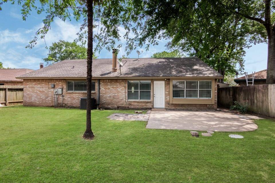 This is a single-story brick home with a spacious lawn, a concrete patio area, and mature trees providing shade.