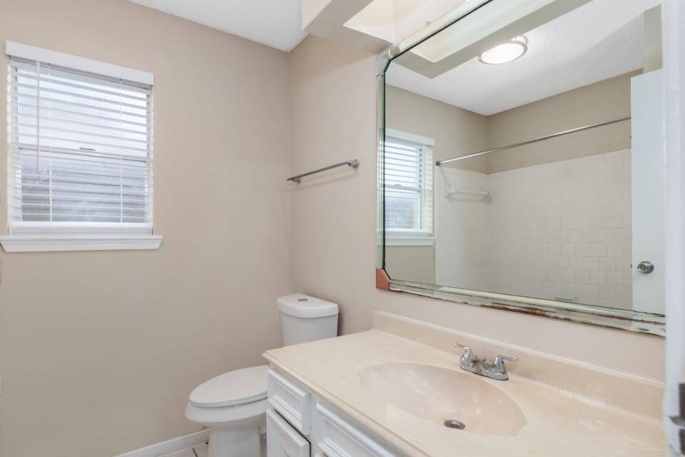Bright bathroom featuring a large mirror, beige walls, and a window with blinds. It includes a toilet and a vanity with a single sink.