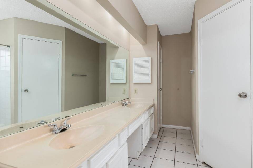 This bathroom features a long countertop with dual sinks, a large mirror, neutral-colored walls, and tiled flooring. It has ample storage with cabinets underneath and a clean, bright appearance.