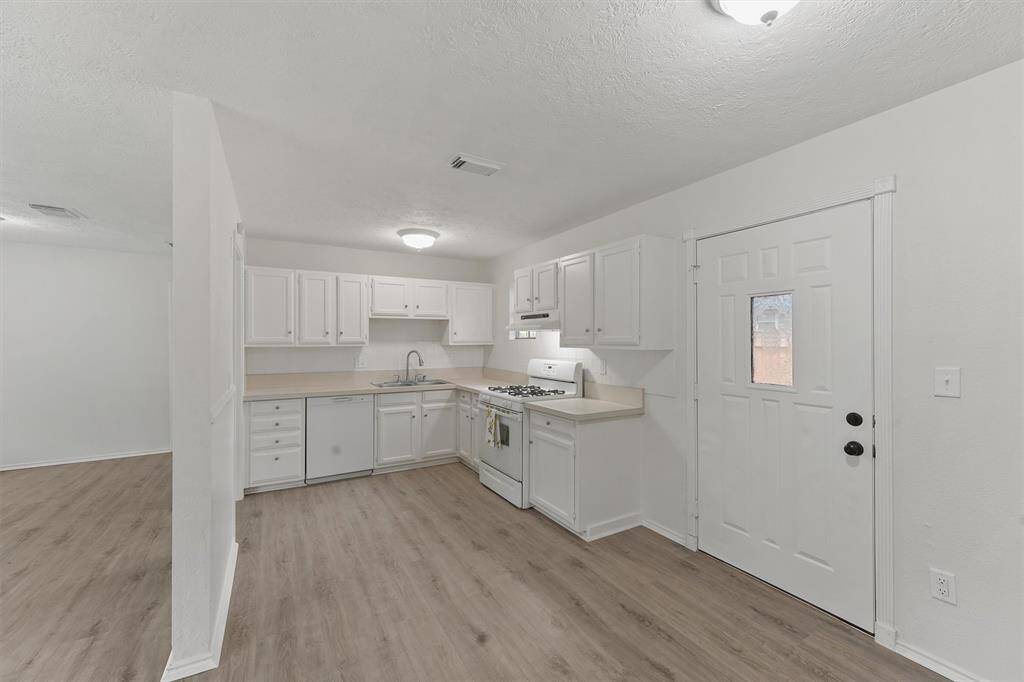 Kitchen with white cabinetry, light countertops, and wood-style flooring. It includes a gas stove, and ample storage space, with a nearby door providing outdoor access.