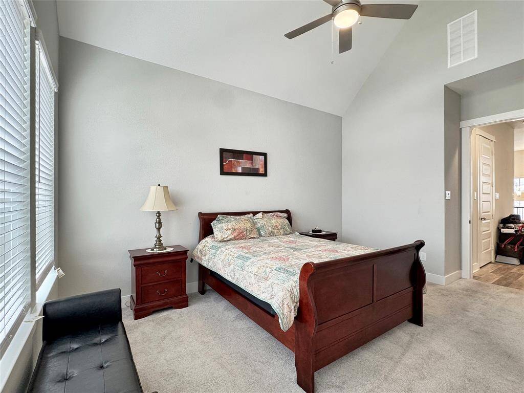 The primary bedroom features a vaulted ceiling with a fan, creating an open and airy feel. It has large windows that provide ample natural light, neutral walls, and cozy carpeting.