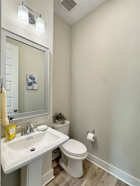 Compact, modern half-bathroom featuring a pedestal sink, a toilet, and a wall-mounted mirror. Light-color walls, wood-style flooring, and a simple light fixture above the sink, create a clean and inviting space.