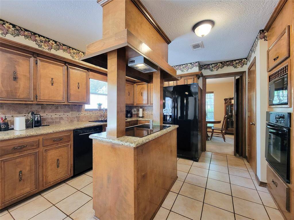 This kitchen features classic wooden cabinetry with granite countertops, a central island, and modern appliances. The space is accented with floral wallpaper trim and has tiled flooring, creating a warm and inviting atmosphere. Natural light enters through a window above the sink, enhancing the cozy feel.