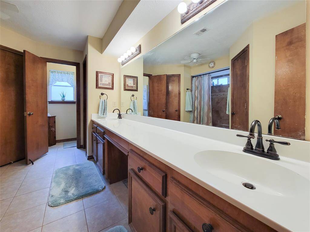 This spacious bathroom features a long vanity with dual sinks, ample storage, and a large mirror. It has warm wood accents, a tiled floor, and a separate area for the shower and toilet, creating a comfortable and functional space.