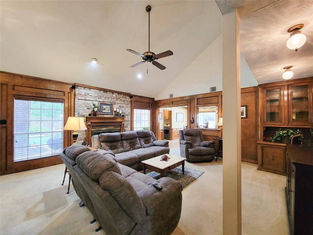 Spacious living room with vaulted ceilings, featuring a cozy brick fireplace, large windows for natural light, and elegant wood paneling. Equipped with plush seating, ceiling fan, and built-in cabinetry, it offers a warm, inviting atmosphere.