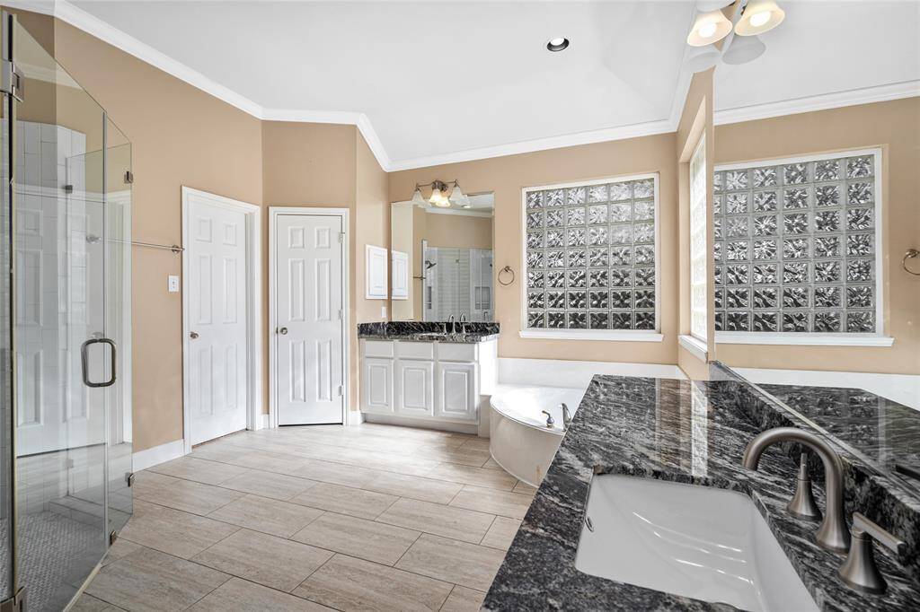 The luxurious primary bathroom is a true retreat, featuring a garden jetted tub positioned beneath two glass brick windows, offering a serene, spa-like atmosphere. His and Her vanities with sleek frameless mirrors provide ample space and modern elegance, while the spacious glass shower adds a touch of sophistication and convenience.