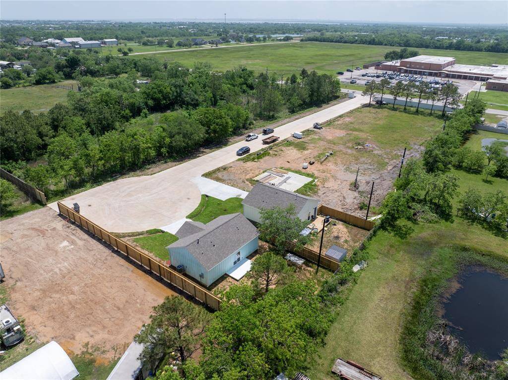 This aerial photo showcases a newly built home on a spacious lot, surrounded by greenery, with a wooden fence. It's situated in a quiet area with a few neighboring houses and open fields, and nearby elementary school offering a peaceful setting.