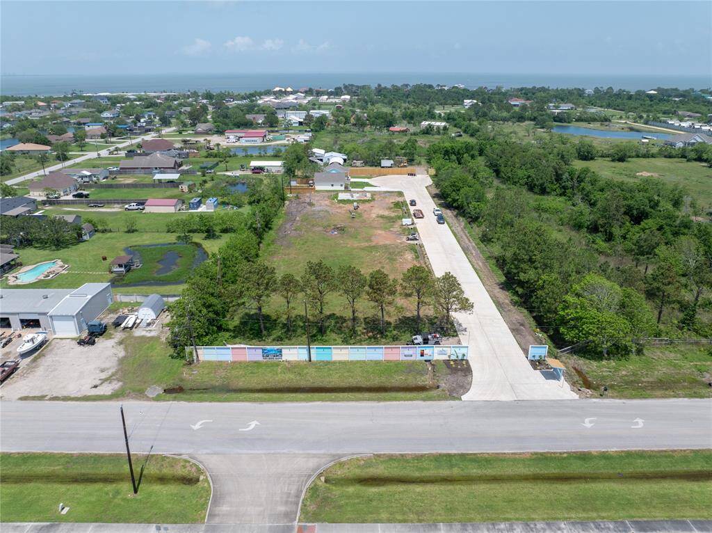 This aerial photo showcases a cleared lot in a suburban neighborhood, bordered by trees and accessible via a paved road. The area is residential with nearby homes and open green spaces, offering a serene setting with potential water views in the distance.