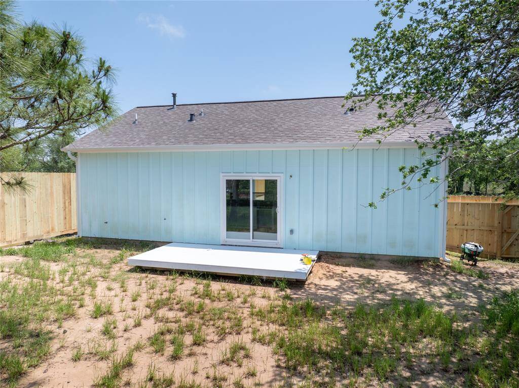 This photo shows the rear view of a house with light blue siding, a small patio, and sliding glass doors. It's surrounded by a partially grassed yard with partial wooden fencing and some trees, offering a private outdoor space.