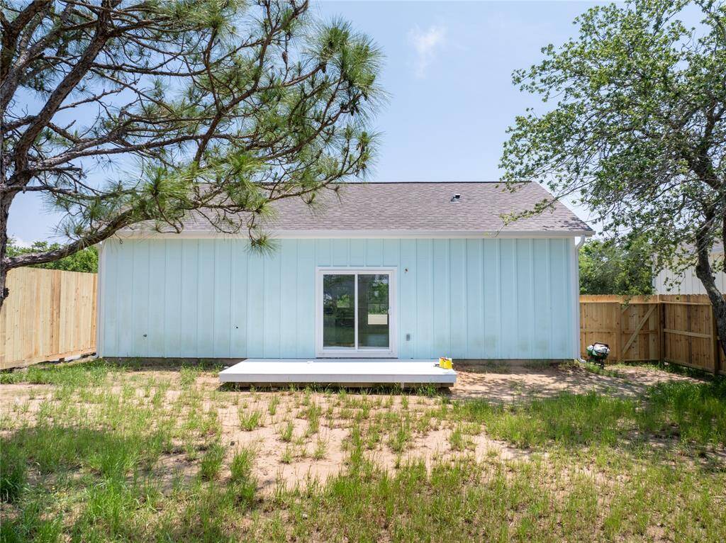 This photo shows the backyard of a house with a light blue exterior, featuring a small deck and sliding glass doors. The yard is partially fenced, with some grass and trees providing shade.