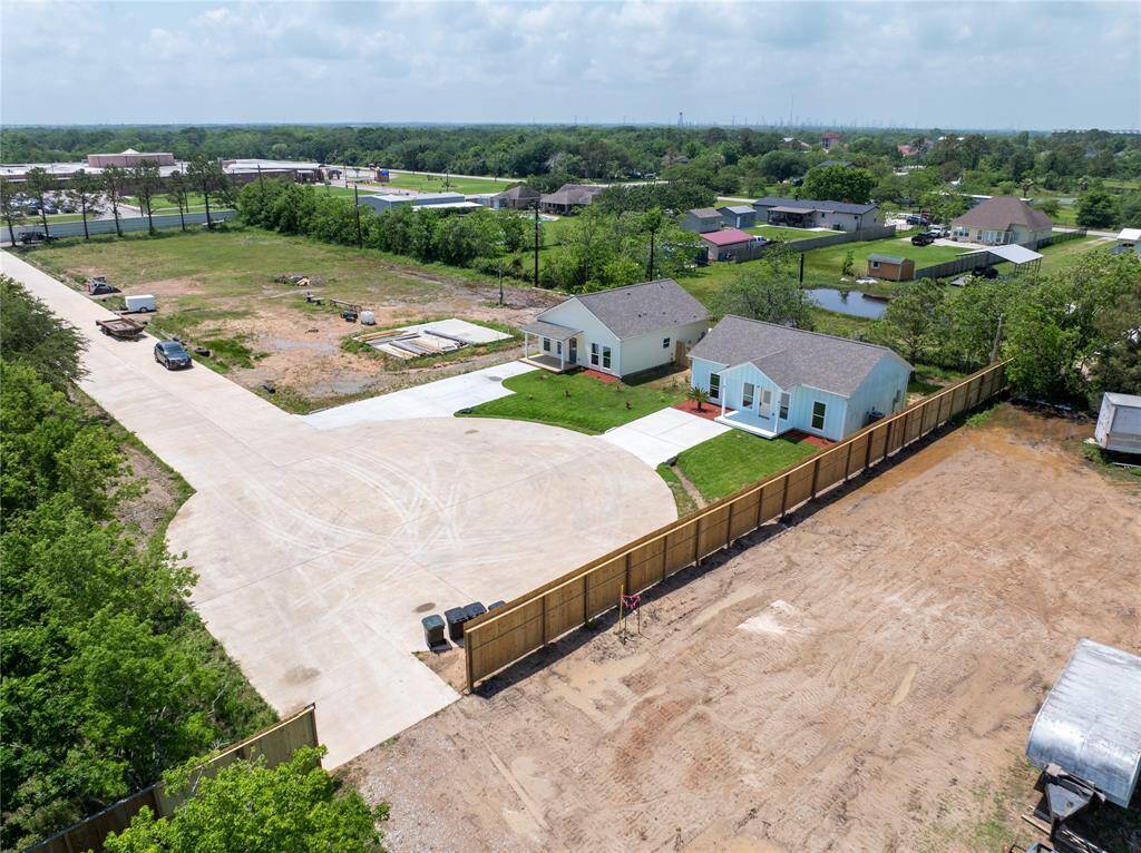 This aerial photo shows two small, newly built homes with neatly maintained lawns on a spacious lot. The area has a paved driveway and is bordered by a wooden fence. The surroundings are open with greenery and other homes nearby, offering a suburban feel.