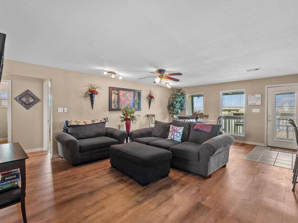 This photo showcases a cozy living room with warm wood flooring and neutral walls. It features a comfortable dark gray sofa set with colorful cushions, a ceiling fan, and ample natural light from multiple windows. The space is accented with vibrant wall art and decorative plants, creating a welcoming atmosphere.