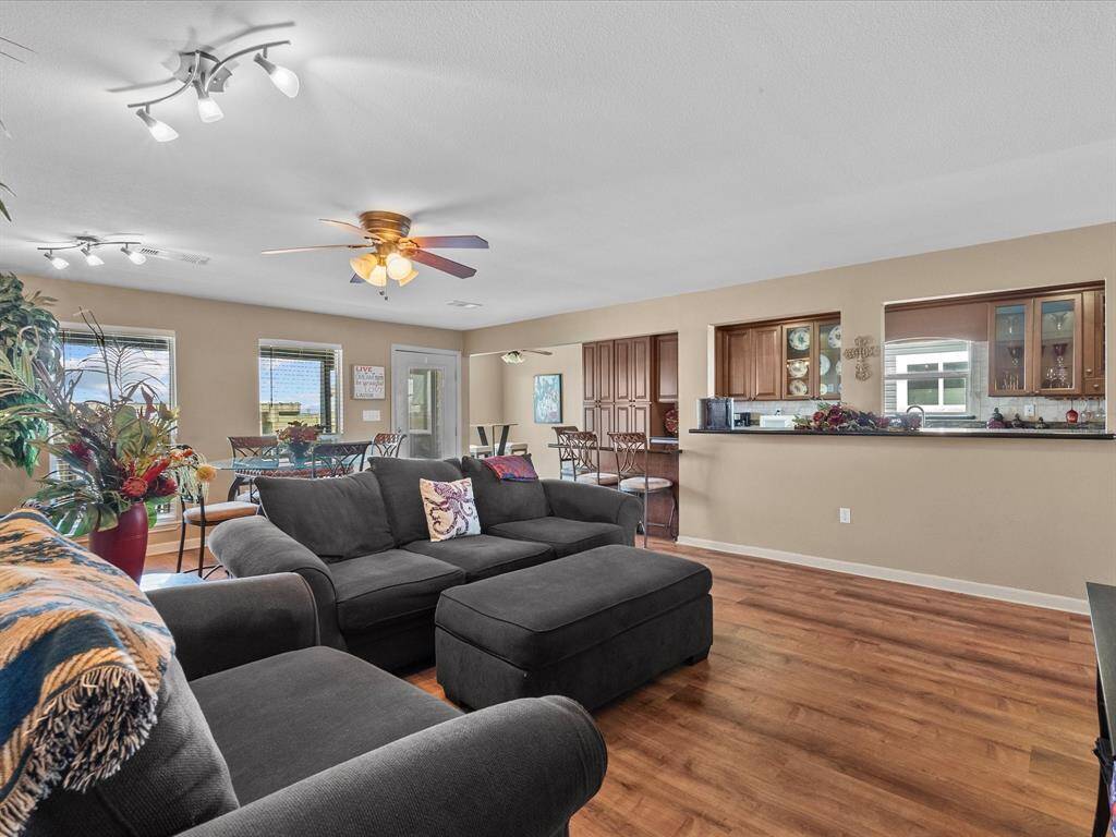 This inviting open-plan living area features warm wood flooring, neutral walls, and ample natural light. It includes a cozy sitting area with a large sectional, a dining space with views, and a kitchen with a convenient breakfast bar. Modern lighting and ceiling fans enhance the ambiance.