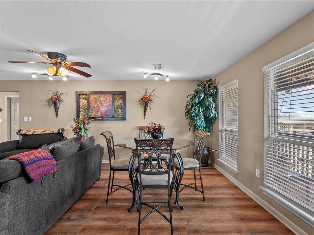 This inviting living and dining area features warm wood flooring and abundant natural light from large windows. The space is tastefully decorated with vibrant artwork, plants, and a cozy seating area complemented by a stylish glass dining table.