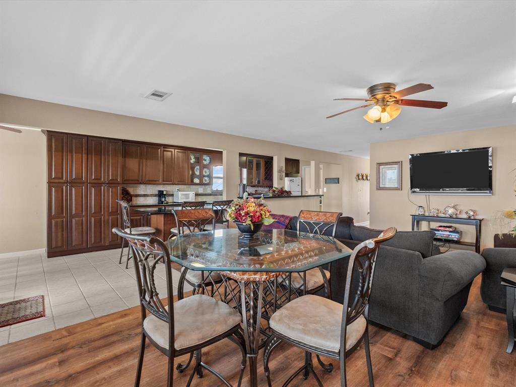 Open-concept living area featuring a spacious kitchen with dark wood cabinets, a breakfast bar, and modern appliances. The dining area has a glass table with metal chairs, leading into a cozy living room with a mounted TV and ceiling fan. The space is bright and inviting, ideal for entertaining.