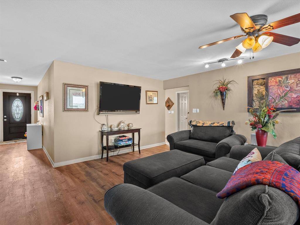 Cozy living room with hardwood floors, neutral walls, and a ceiling fan. Features a welcoming entryway, wall-mounted TV, and comfortable seating. Decor includes vibrant artwork and plants, creating a warm, inviting atmosphere.