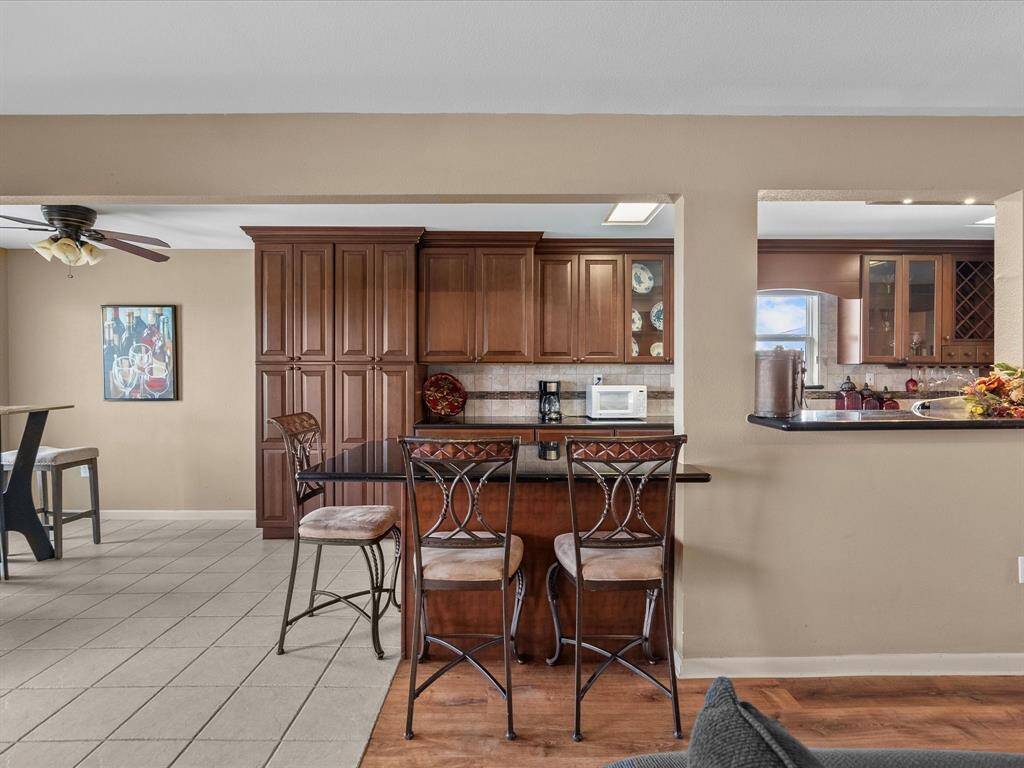 This photo features a cozy kitchen with rich wooden cabinetry and a breakfast bar with two stools. The space is open to a dining area with a ceiling fan and tile flooring, creating a warm and inviting atmosphere perfect for entertaining.