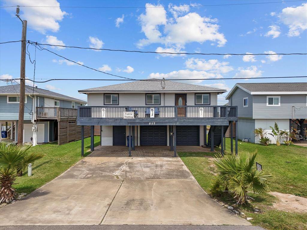 Elevated two-story home with a spacious balcony, two-car garage, and a wide driveway. The exterior features a modern design with palm trees and a well-maintained lawn.