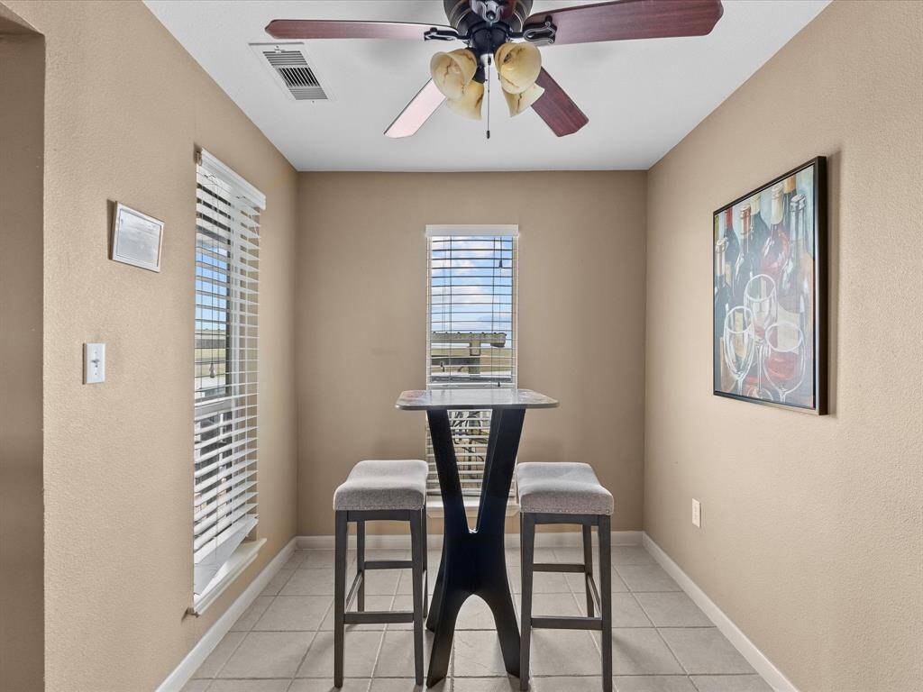 Cozy dining nook with neutral walls, featuring a modern high-top table and two upholstered stools. Natural light streams through the window with blinds, and a ceiling fan adds comfort. A decorative painting adds a touch of elegance.