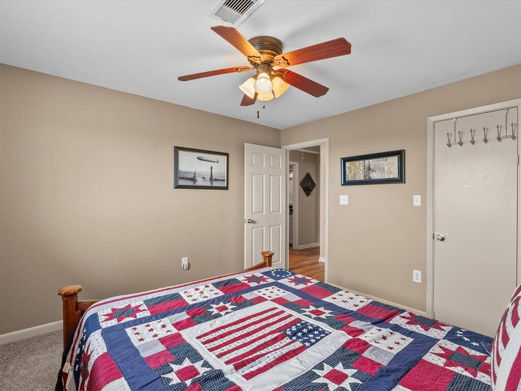 Cozy bedroom with neutral walls, a ceiling fan, and carpeted flooring. Features a decorative bedspread and two wall hangings, creating a warm and inviting atmosphere.