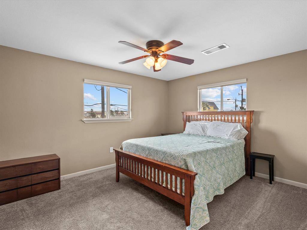 This bedroom features neutral walls and carpet, a ceiling fan, and ample natural light from two windows. It includes a wooden bed frame and a small dresser, offering a cozy and functional space.