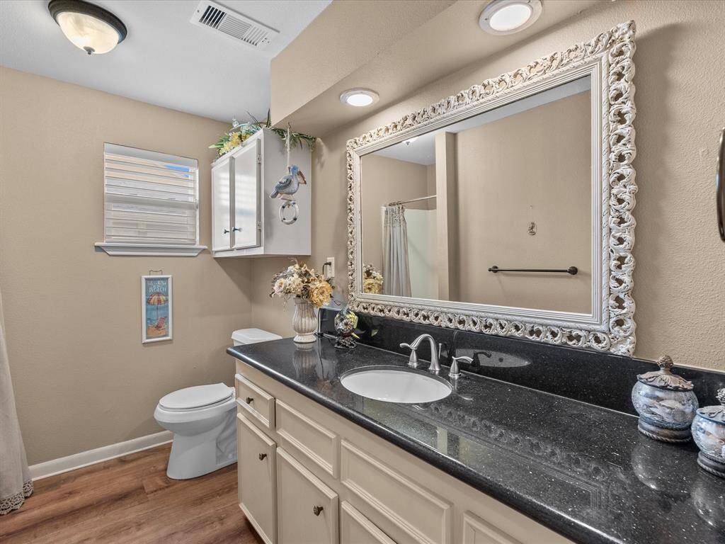 This bathroom features a modern design with neutral tones and wood flooring. It includes a sleek black countertop with a white sink, a large ornate mirror, and ample cabinet storage. The space is well-lit with both ceiling and wall fixtures, and it has a shower area with a curtain. Decor includes floral arrangements and a small wall cabinet for additional storage.