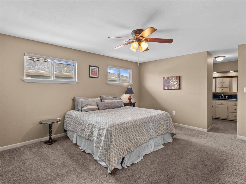 This spacious bedroom features neutral walls, a ceiling fan, and carpeted flooring. It includes a large bed with side tables and lamps, and two small windows for natural light. An ensuite bathroom is visible, adding convenience.