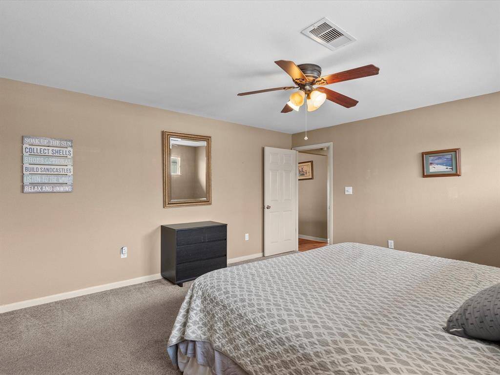 This bedroom features neutral walls, carpeted flooring, and a ceiling fan with lights. It includes a large bed, a small dresser, and wall art, creating a cozy and inviting space.