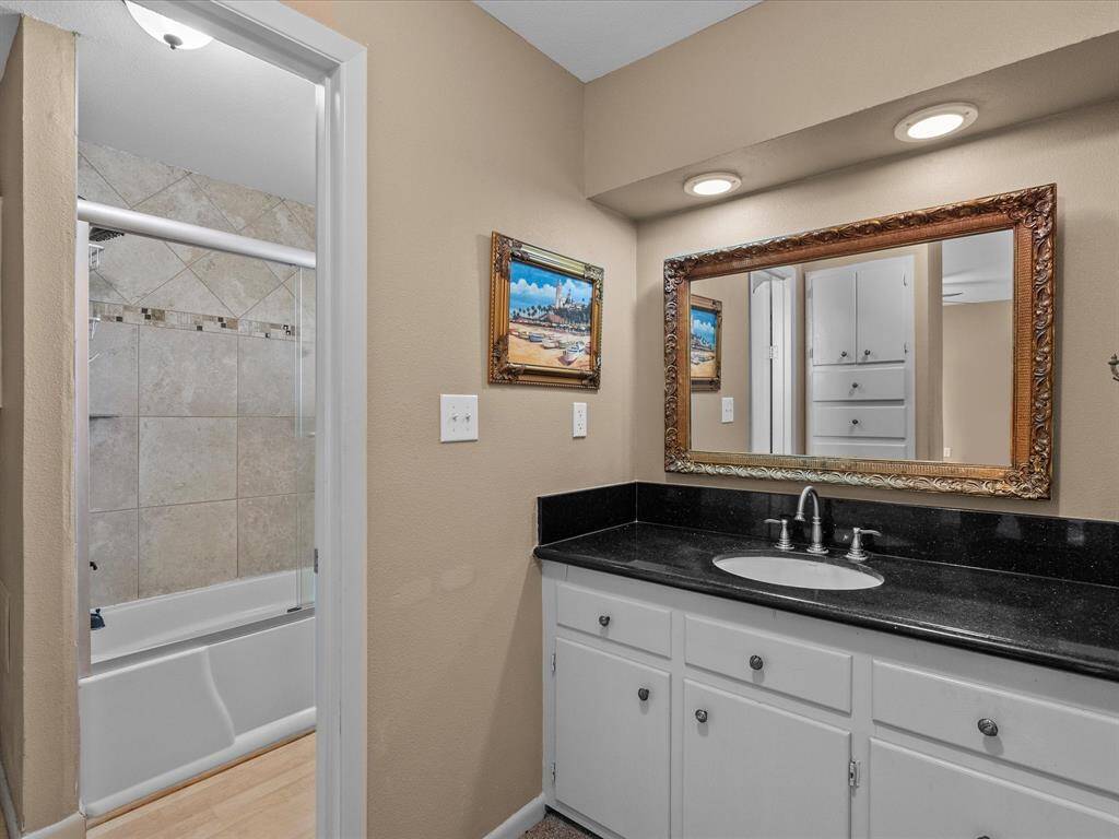 This bathroom features a sleek black countertop with a single sink, white cabinetry, and a large decorative mirror. It includes a shower-tub combo with tiled walls and recessed lighting, offering a clean and stylish space.