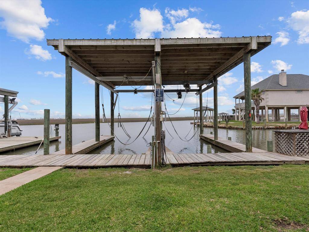 This photo shows a waterfront property with a covered boat dock. It features a wooden structure with water access, ideal for boating enthusiasts. A house is visible in the background, surrounded by grass and a clear blue sky. The home is minutes from the Gulf.
