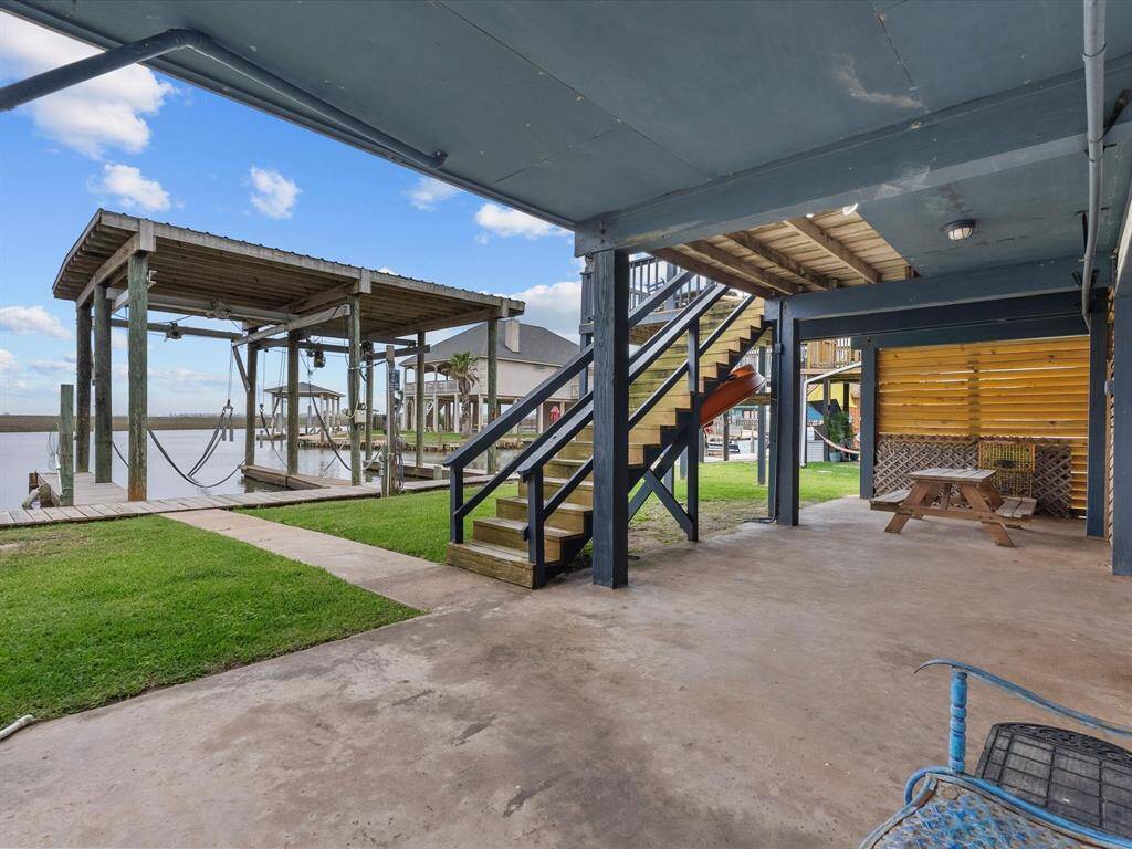 This photo showcases a covered patio area with outdoor seating and a wooden staircase leading to an upper level. The space opens to a grassy area and a waterfront view, complete with a dock and boat lift, ideal for boating and fishing enthusiasts.