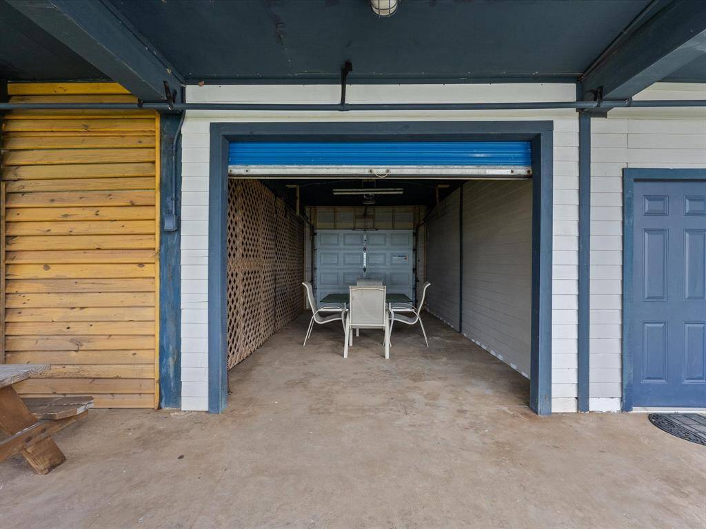 This photo shows a partially enclosed garage space with a table and chairs set inside. It features a mix of wood and painted surfaces, with a closed garage door at the back and a side door to the right. The area is versatile, suitable for storage or a workspace.