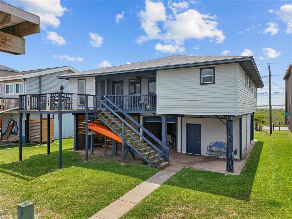 Elevated coastal home with a spacious deck, featuring a sturdy staircase leading to the main entrance. The property includes a covered patio area beneath the house, ideal for outdoor activities, and is surrounded by a well-maintained lawn.