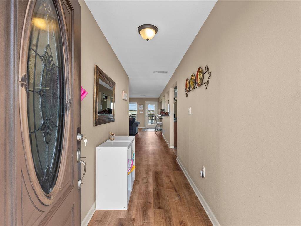 This welcoming entryway features warm wood flooring and neutral walls, leading to an open living space. Decorative accents and a large mirror enhance the inviting atmosphere, while natural light filters in from the far end, creating a bright and airy feel.