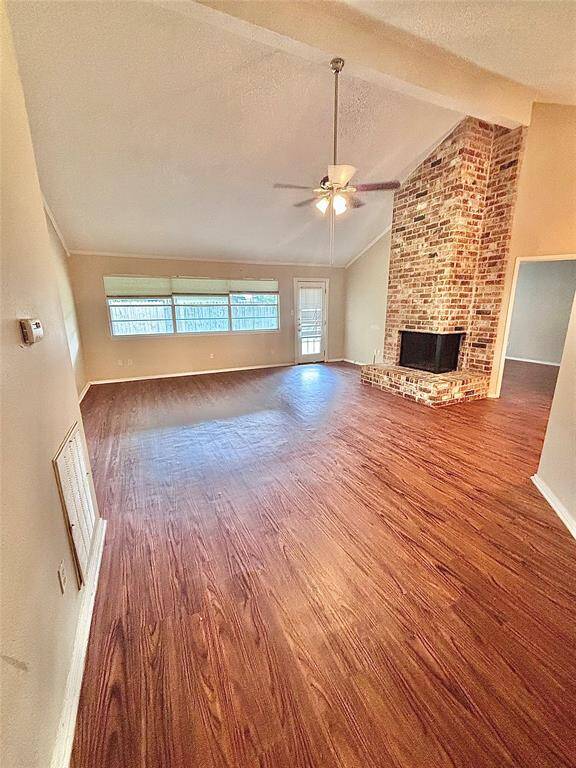 Spacious living room with high ceilings, featuring a charming brick fireplace. It has rich wood flooring, ample natural light from large windows, and ceiling fan for comfort.