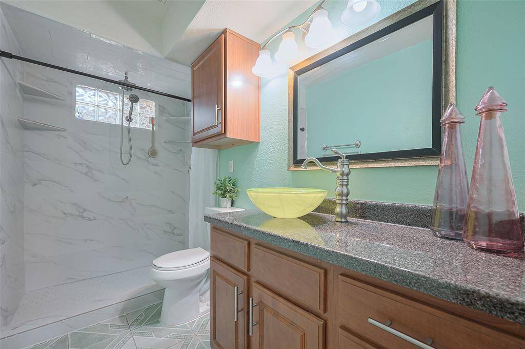 The home features one full bath and a half bathroom. The full bath includes a vanity with a granite-look laminate countertop, a vessel sink, and an updated faucet. A decorative framed mirror, updated overhead lighting, and a commode with an over-commode storage cabinet add stylish touches. The 2024 remodeled walk-in shower boasts dual showerheads, a glass block privacy window, and multiple ledges for shampoo and soap, combining both luxury and functionality.