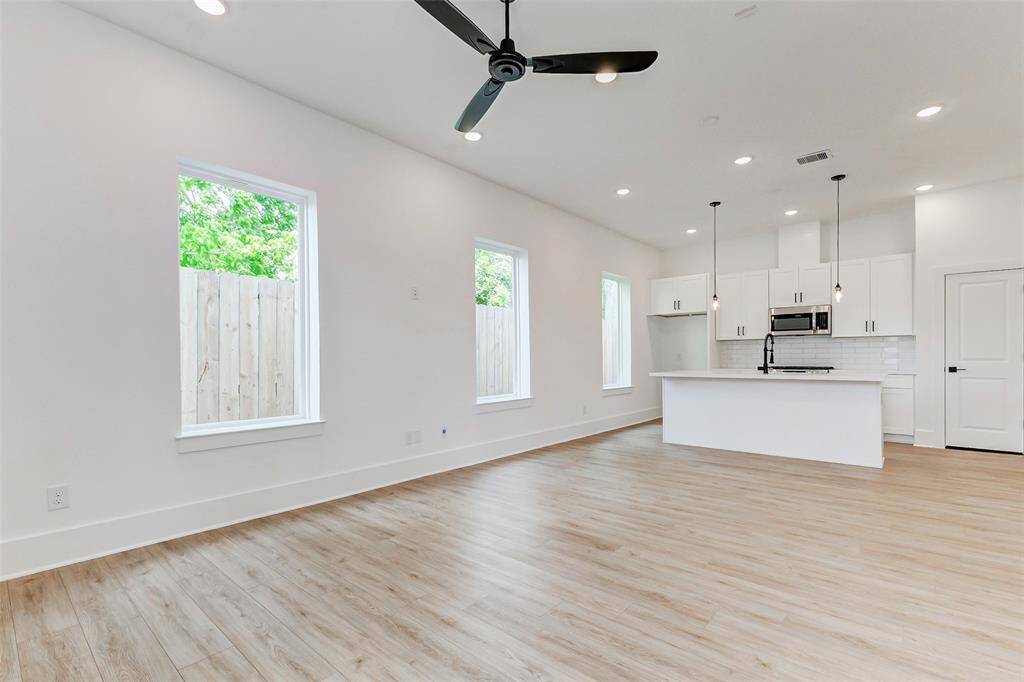 Bright, open-concept living area with light wood flooring and a modern kitchen. The kitchen features white cabinetry, a large island, and stainless steel appliances. Ample natural light streams through multiple windows, and a ceiling fan adds comfort to the space.