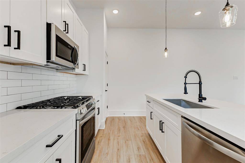 Modern kitchen featuring sleek white cabinetry, stainless steel appliances, and a stylish subway tile backsplash. The space includes a gas stove, built-in microwave, elegant pendant lighting, and a contemporary faucet over a large sink, all set against a light  floor.