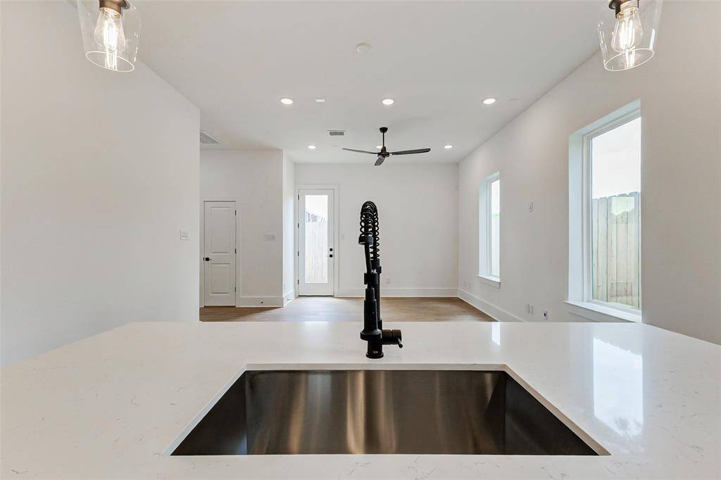 Open-concept living area with a bright, airy feel. It features a sleek kitchen island with a contemporary faucet, recessed lighting, large windows for natural light, and a ceiling fan. The white walls and light wood flooring enhance the spaciousness.