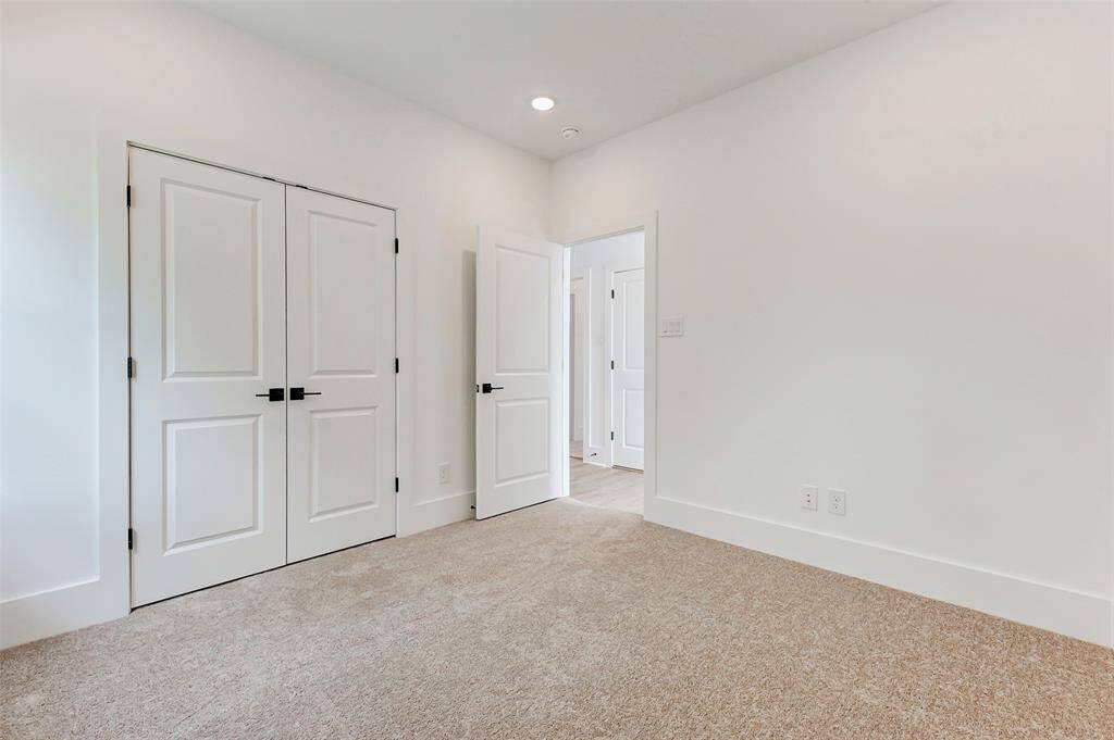 This room features clean white walls, soft carpet flooring, and recessed lighting. It includes a double-door closet and a doorway leading to a hallway, offering a bright and inviting space ideal for a bedroom or office.