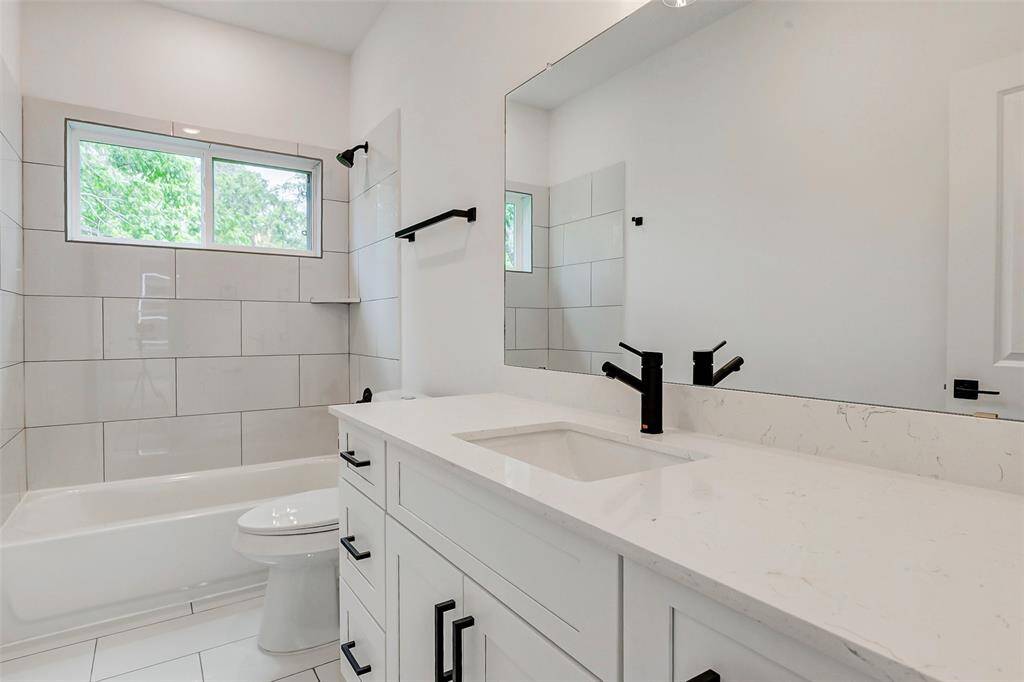 This bathroom features a modern design with a white vanity, black fixtures, and a large mirror. It includes a tiled shower-tub combo with a window for natural light, creating a bright and airy space.