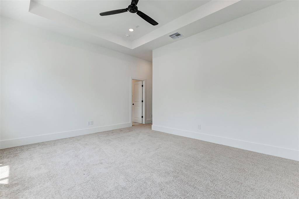 This spacious room features neutral carpeting, crisp white walls, recessed lighting, and a modern ceiling fan. The space offers ample natural light and leads to an adjoining area through a door.