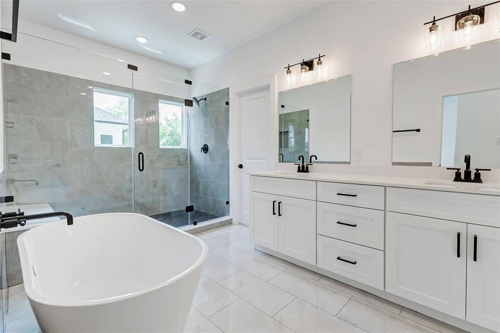 Modern and spacious bathroom featuring a large walk-in shower with glass doors, a freestanding bathtub, and a double vanity with ample storage. The room is bright with white cabinetry and sleek black fixtures, creating a clean and elegant look.
