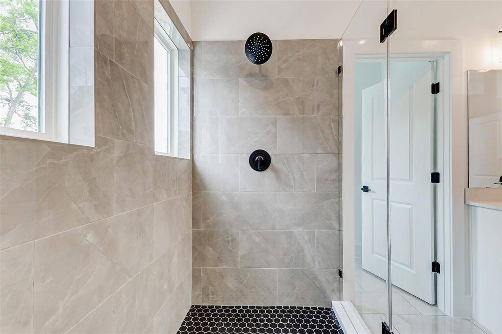 Experience luxury in this modern bathroom with a spacious walk-in shower, sleek gray tiles, and stylish black fixtures. Abundant natural light pours through dual windows, enhancing its contemporary charm.