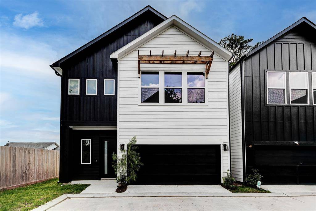 Gated community. Modern two-story home with contrasting black and white siding, featuring a covered entrance and large windows. The driveway leads to a double garage.