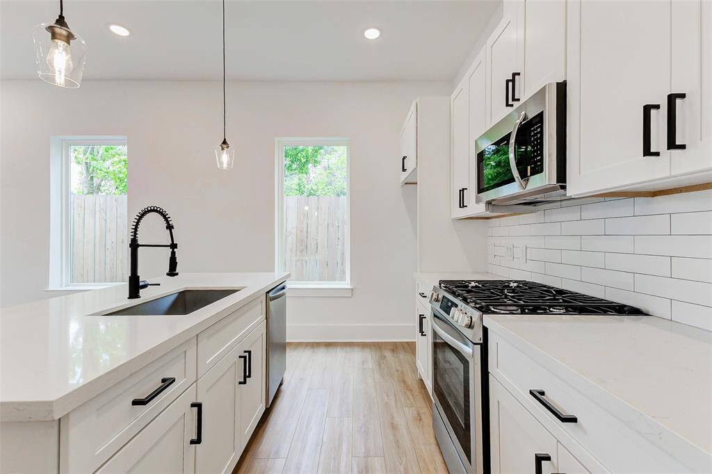 This kitchen features a modern design with white cabinetry, stainless steel appliances, and a sleek gas stove. It has large windows for natural light, elegant pendant lighting, and a spacious island with a stylish black faucet, all set on light LVP flooring. Refrigerator included