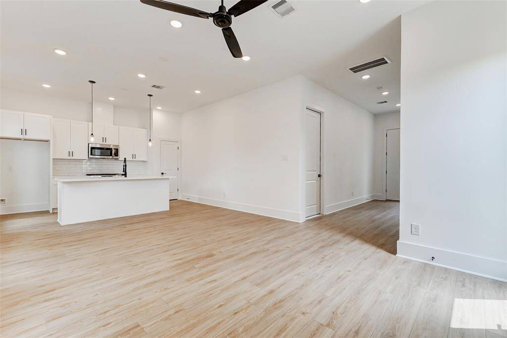 Modern, open-concept living space with light wood flooring and a minimalist kitchen. The kitchen features white cabinetry, a central island with pendant lighting, and stainless steel appliances. The area is bright and airy, with recessed lighting and a ceiling fan.