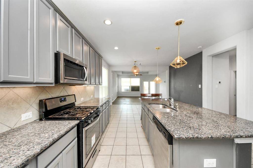 This spacious kitchen features modern gray cabinets, granite countertops, and stainless steel appliances. It includes a large island with a double sink, pendant lighting, and an open layout leading to the dining area, perfect for entertaining.