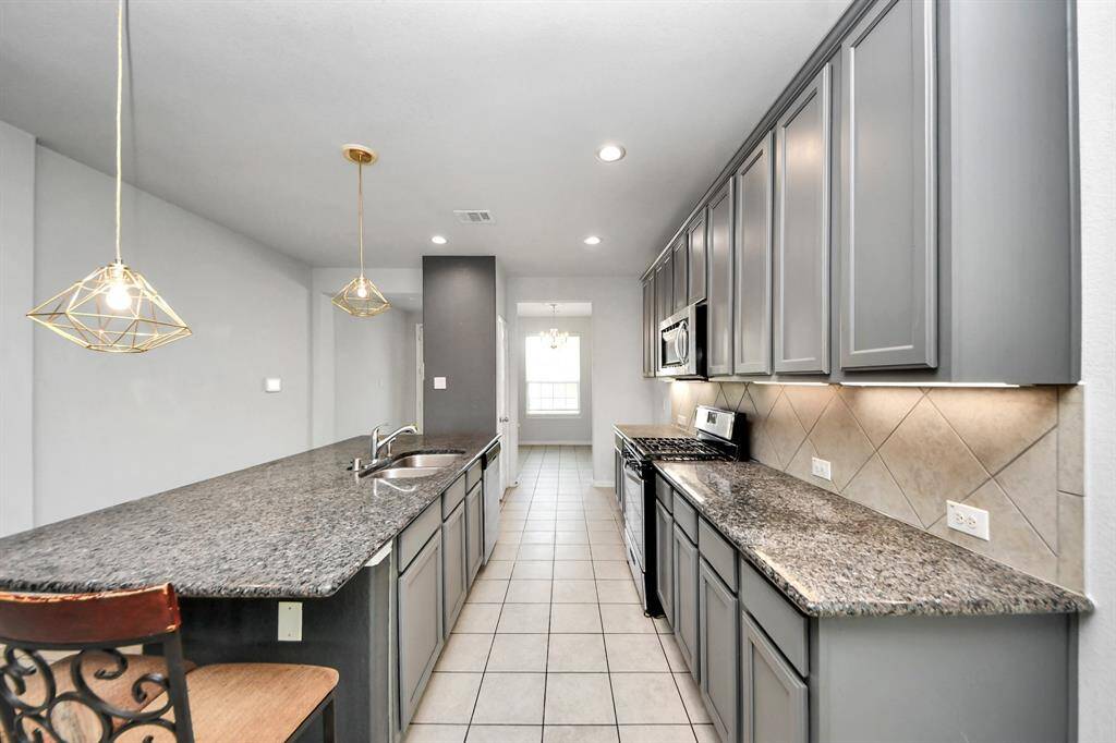 This kitchen features sleek gray cabinetry, granite countertops, and modern pendant lighting. It's equipped with stainless steel appliances and has a spacious layout with a tiled floor and ample storage.