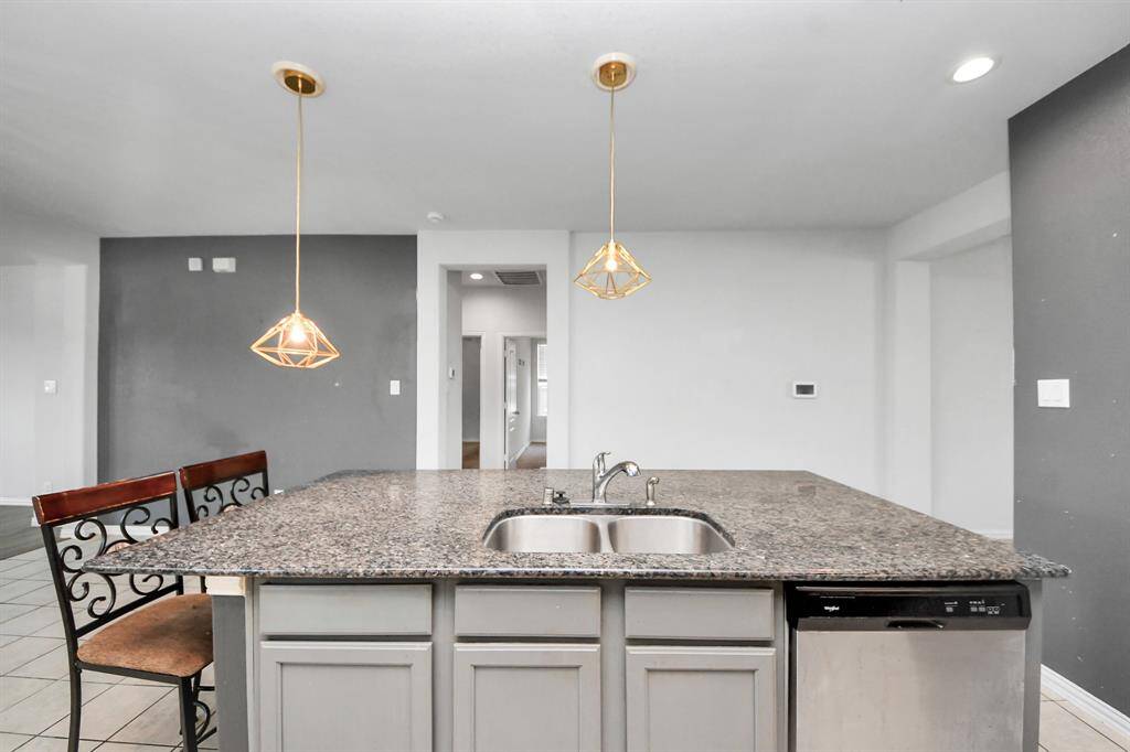This photo showcases a modern kitchen with a granite-topped island, dual sink, and stainless steel dishwasher. Elegant pendant lights hang above, and the space features a neutral color palette with a gray accent wall.